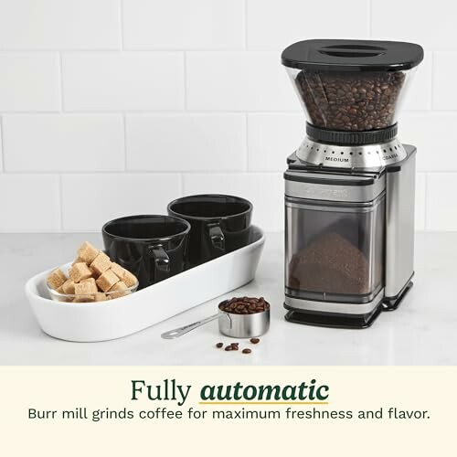 Coffee grinder with coffee beans, two black cups, and sugar cubes.