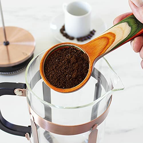 A hand holding a colorful scoop with ground coffee over a French press, showcasing the scoop's functionality and style.